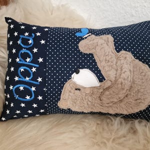 Könnte beinhalten: Ein rechteckiges Kissen in Marineblau mit weißen Punkten und Sternen. Das Wort "Oppa" ist blau auf der linken Seite gestickt. Auf der rechten Seite befindet sich eine Applikation eines hellbraunen Teddybären mit einem blauen Schmetterling auf dem Kopf.
