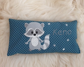 Puede incluir: Un cojín rectangular de color verde azulado con un patrón de lunares blancos y un mapache gris bordado. El nombre Keno está cosido con hilo gris en el lado derecho, acompañado de pequeñas estrellas blancas bordadas sobre la tela.