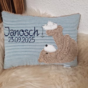 Könnte beinhalten: Ein rechteckiges Kissen mit blau-weißem Querstreifenmuster. Es ist mit dem Namen Janosch, dem Datum 23.09.2025 sowie einer Applikation aus braunem Plüsch bestickt, die einen Bären mit einem weißen Papierboot und einem Anker zeigt.