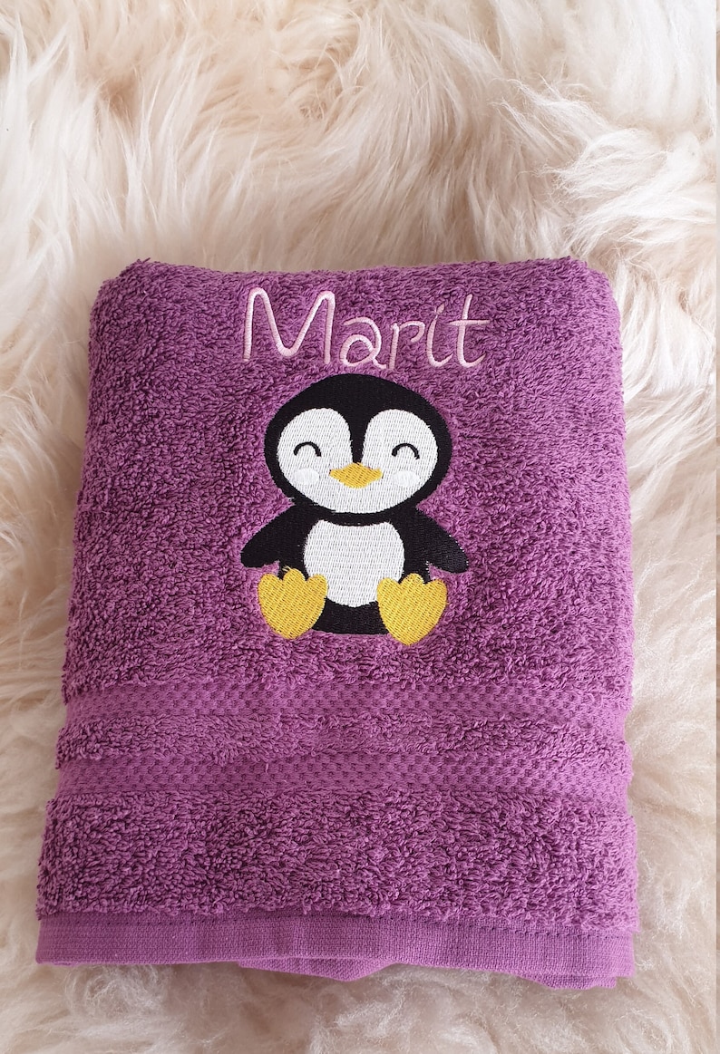 Puede incluir: Una toalla morada doblada con el nombre "Marit" bordado en escritura blanca. Un ping&uuml;ino de dibujos animados con cuerpo negro, vientre blanco, pies amarillos y pico amarillo est&aacute; bordado debajo del nombre. La toalla est&aacute; hecha de un material suave y absorbente.