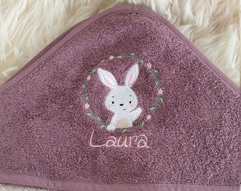 May include: A dusty rose hooded towel featuring an embroidered design of a white rabbit inside a floral wreath. The name Laura is embroidered in pink thread below the rabbit. The towel has a textured terry cloth finish and a smooth fabric trim.
