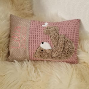 Könnte beinhalten: Ein rechteckiges Kissen mit einer beigen Seite mit weißen Punkten, bestickt mit dem Namen Lilla und dem Datum 24.06.2026 in Pink, sowie einer rosafarbenen Waffelstruktur-Seite mit einer Applikation eines braunen Plüschbären mit Schmetterling.