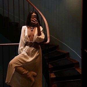 May include: A woman in a white maxi dress with a plunging neckline and a brown belt poses on a staircase. She is holding a phone up to her face and taking a selfie.