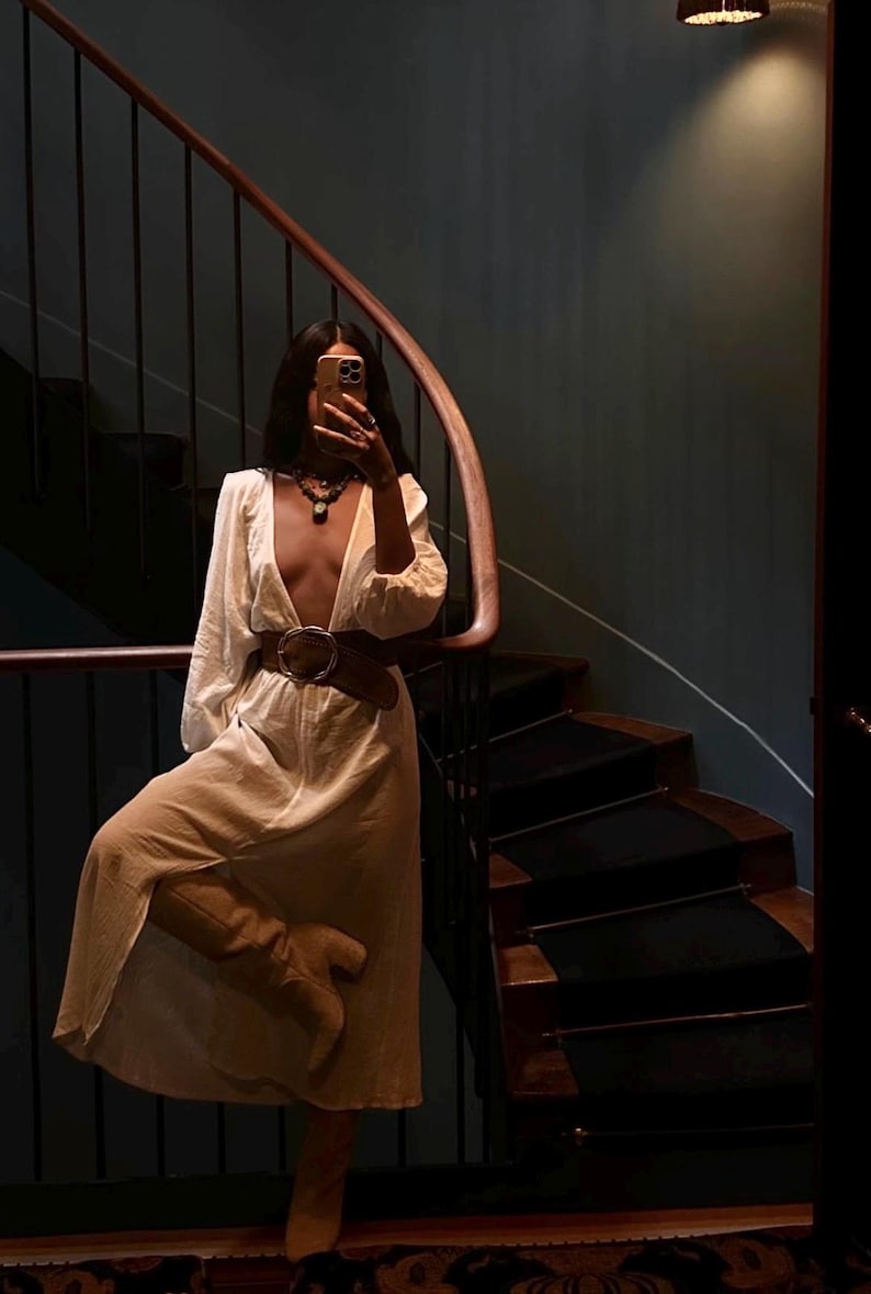 May include: A woman in a white maxi dress with a plunging neckline and a brown belt poses on a staircase. She is holding a phone up to her face and taking a selfie.