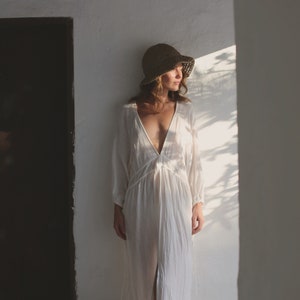 May include: A woman wearing a white linen dress and a straw hat stands in a doorway. She is holding a basket of lemons.