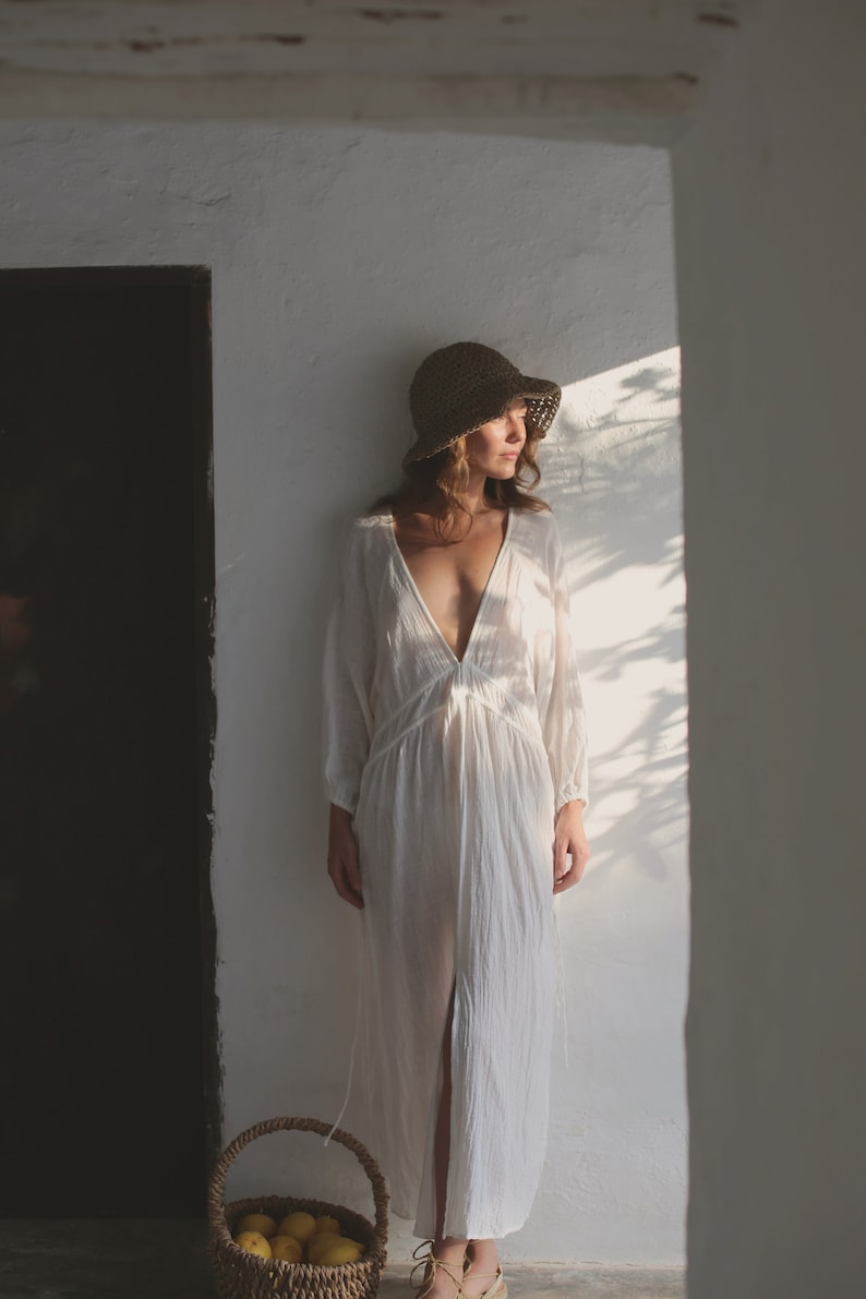 May include: A woman wearing a white linen dress and a straw hat stands in a doorway. She is holding a basket of lemons.