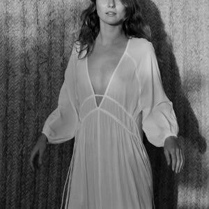 May include: A woman wearing a white, long-sleeved, loose-fitting dress with a deep V-neckline and a tie at the waist. The dress has a sheer, flowing fabric and is accented with delicate, thin ties that hang from the waist.