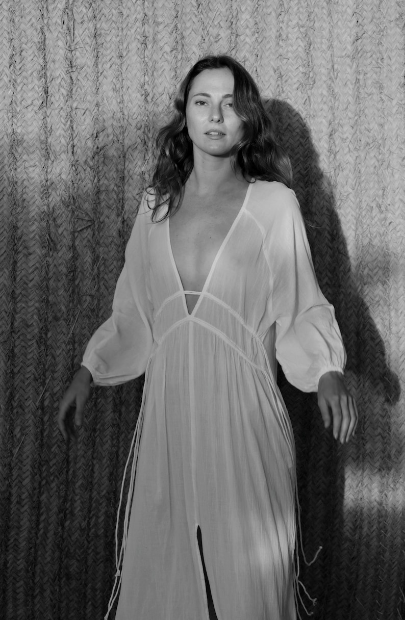 May include: A woman wearing a white, long-sleeved, loose-fitting dress with a deep V-neckline and a tie at the waist. The dress has a sheer, flowing fabric and is accented with delicate, thin ties that hang from the waist.
