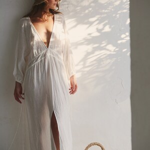 May include: A woman wearing a white linen dress and a straw hat stands in front of a white wall. She is holding a wicker basket filled with lemons.