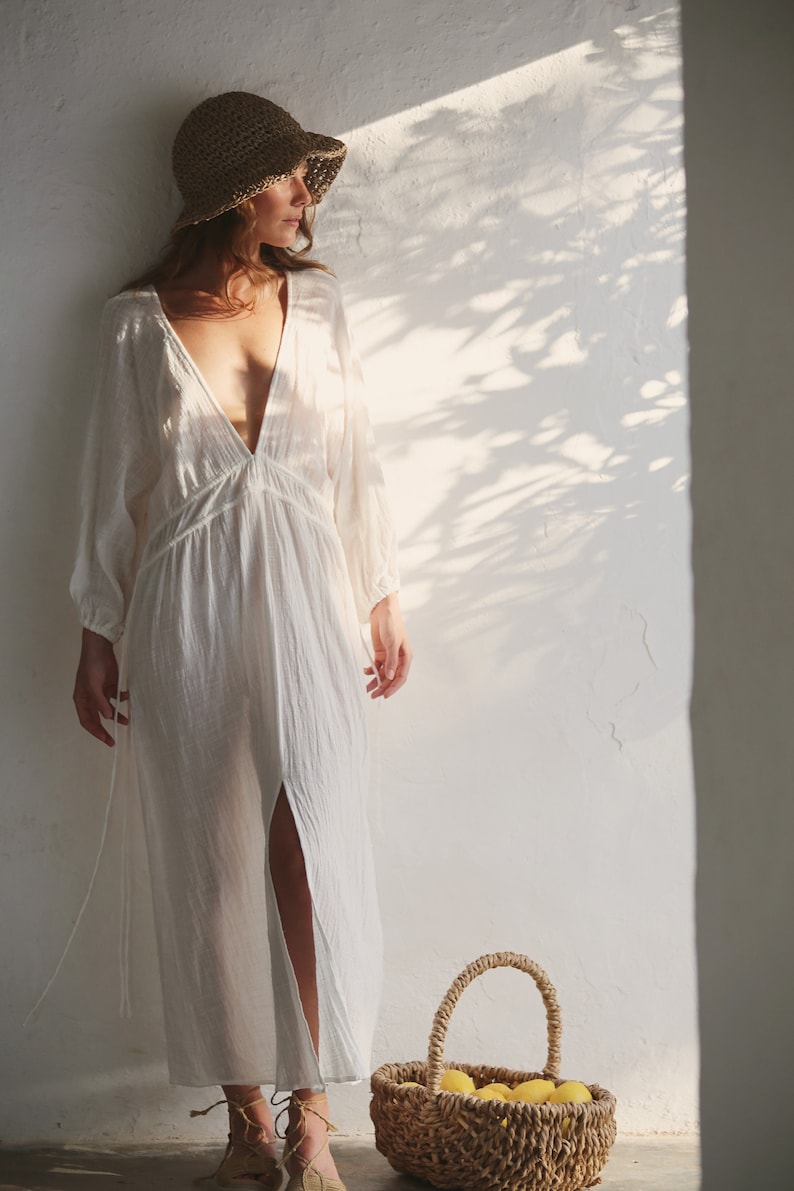 May include: A woman wearing a white linen dress and a straw hat stands in front of a white wall. She is holding a wicker basket filled with lemons.