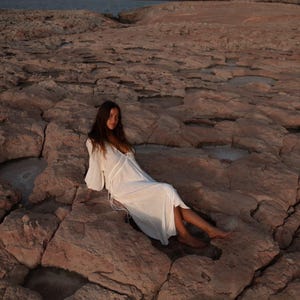 May include: A woman in a white maxi dress sits on a rocky beach. The dress has a V-neckline and long sleeves. The woman's hair is long and brown.