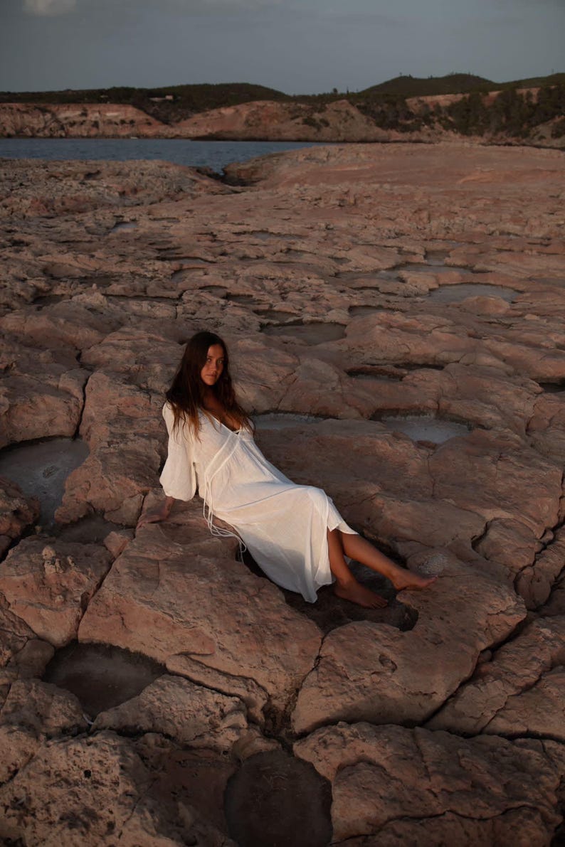 May include: A woman in a white maxi dress sits on a rocky beach. The dress has a V-neckline and long sleeves. The woman's hair is long and brown.