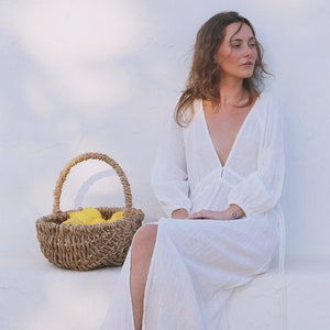 May include: A woman wearing a white maxi dress with a plunging neckline and long sleeves sits against a white wall. She is holding a wicker basket filled with lemons.