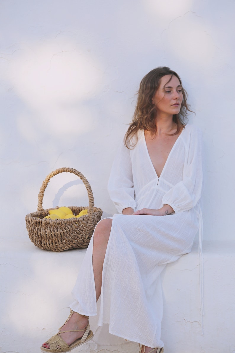 May include: A woman wearing a white maxi dress with a plunging neckline and long sleeves sits against a white wall. She is holding a wicker basket filled with lemons.