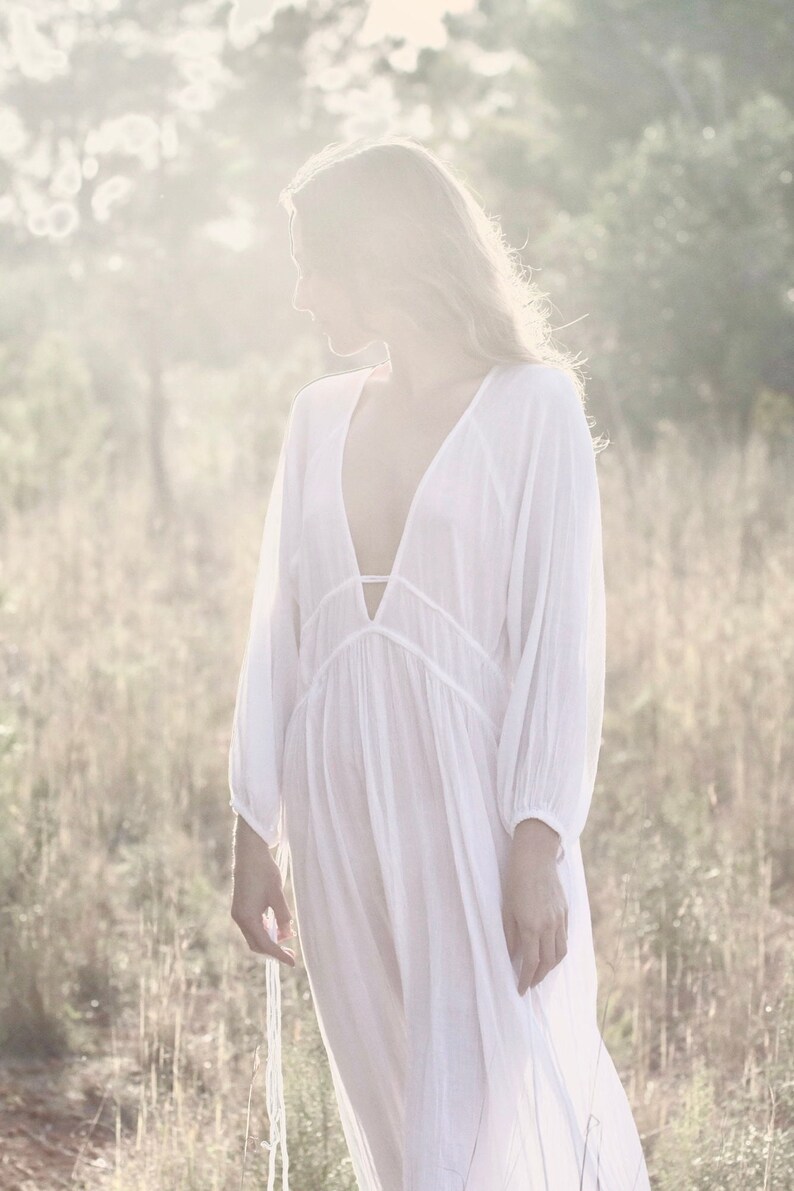 May include: A woman wearing a white, flowing, long-sleeved dress with a deep V-neckline. The dress has a gathered skirt and is made of a lightweight fabric. The woman is standing in a field of tall grass, with the sun shining behind her.