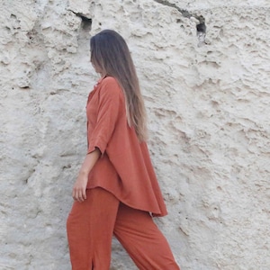 May include: Rust-colored linen outfit, including a button-down shirt with three-quarter sleeves and matching pants. The model is standing in front of a textured, light-colored rock formation. The outfit has a relaxed fit.