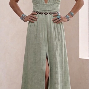 May include: A sage green maxi dress with a plunging neckline and a high slit. The dress features thin straps and a cinched waist. Accessories include turquoise jewelry and a beaded belt. The dress is made of a textured fabric.
