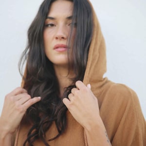 May include: A woman wearing a brown hooded poncho with a drawstring. The poncho has a fringed hem and is made of a soft, textured fabric. The woman has long, dark hair and is looking at the camera.