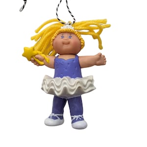 May include: A Cabbage Patch Kids ballerina ornament. The doll has yellow yarn hair, a purple leotard with star accents, a white tutu, and purple pants. It holds a yellow star wand and has a black and white string for hanging.