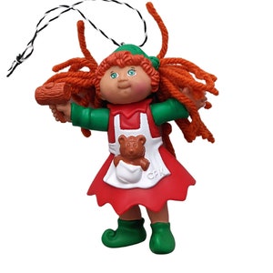May include: A red-haired Cabbage Patch Kid doll dressed as an elf, wearing a green hat, red dress, and white apron. The doll is holding a wooden log and has a teddy bear in its apron pocket. The doll is a Christmas ornament.