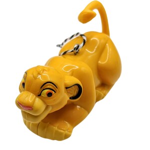 May include: A yellow plastic ornament of Simba from Disney's The Lion King. Simba is lying down with his tail curled up behind him. He has a black nose, black eyes, and a pink tongue.