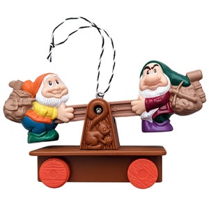 May include: A decorative ornament featuring two dwarf figures balancing a seesaw. One dwarf wears an orange hat and blue jacket, the other a green hat and burgundy jacket. The ornament has a brown base with red wheels and a string for hanging.