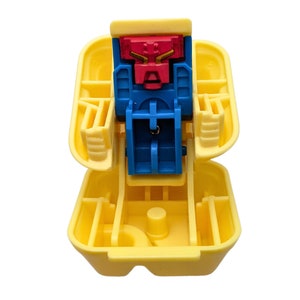 May include: A yellow plastic toy with a hinged lid, revealing a red and blue robot-like figure. The figure has a red head and blue body. The toy is open, showing its internal structure and design.