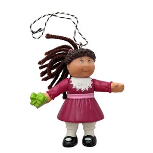 May include: A Cabbage Patch Kid ornament with brown yarn hair, a pink dress with white trim, and a green gift. The ornament has a string for hanging.
