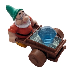 May include: A plastic toy figurine of a dwarf with a green hat and a red and white shirt pushing a brown toy wagon filled with blue gems and white ice.