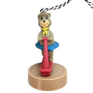 May include: A brown bear wearing a blue skirt and yellow scarf rides a red pogo stick. The bear is a Christmas ornament with a brown base.