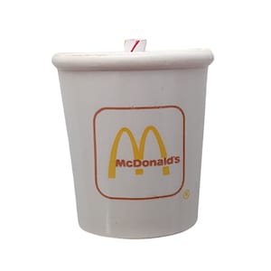 May include: A white McDonald's soda cup with the golden arches logo and the McDonald's name in red. The cup has a red and white striped straw and a brown border around the logo.