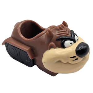May include: A brown Taz-Mania character toy car with black wheels. The car features the Tasmanian Devil's face, with a tan muzzle, black nose, and white eyes. The car has a small opening in the back.