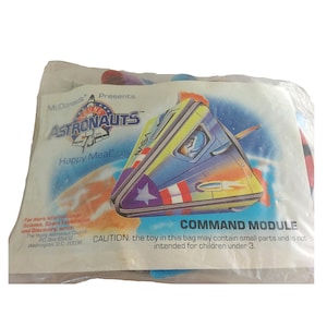 May include: A blue and yellow plastic bag with the words "McDonald's Presents Young Astronauts Happy Meal" and "Command Module" printed on it. The bag contains a toy space shuttle. A warning label reads "CAUTION: the toy in this bag may contain small parts and is not intended for children under 3."