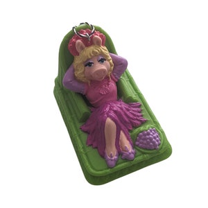 May include: A green plastic figurine of Miss Piggy from The Muppets, wearing a pink dress and a purple flower crown, relaxing on a green plastic lounge chair with a purple grape design.