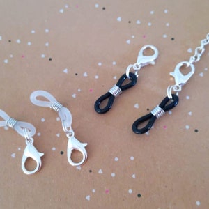 Könnte beinhalten: Brillenkette mit silberfarbenen Metallverschlüssen und -verbindern. Die Brillenhalter haben schwarze und transparente Gummischlaufen zur Aufnahme der Brille. Die Kette ist silberfarben.