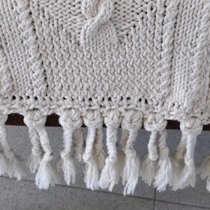 Könnte beinhalten: Eine weiße Häkeldecke mit einem Zopfmuster und Fransenbesatz.