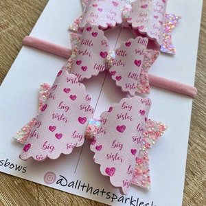 May include: Two pink hair bows with glitter and a white background. The bows are decorated with pink hearts and the words "big sister" and "little sister".