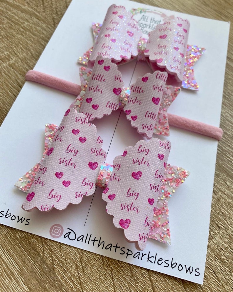 May include: Two pink hair bows with glitter and a white background. The bows are decorated with pink hearts and the words "big sister" and "little sister".