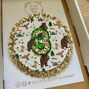 K&ouml;nnte beinhalten: Ein goldener Glitzerkreis mit wei&szlig;em Hintergrund und einer gr&uuml;nen Glitzerzahl 3. Der Kreis hat einen wei&szlig;en Hintergrund mit einem Muster aus braunen und gr&uuml;nen Illustrationen eines Charakters aus dem Buch Wo die wilden Kerle wohnen. Der Kreis hat einen goldenen Glitzerrand. Der Text "All that Sparkles Bows" befindet sich unten im Bild.