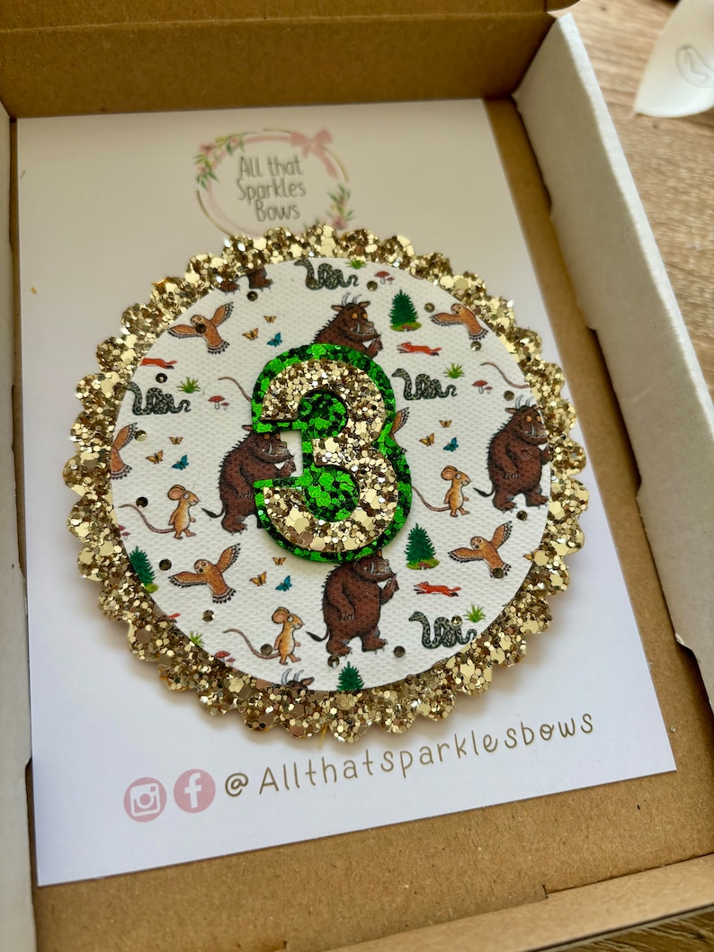 K&ouml;nnte beinhalten: Ein goldener Glitzerkreis mit wei&szlig;em Hintergrund und einer gr&uuml;nen Glitzerzahl 3. Der Kreis hat einen wei&szlig;en Hintergrund mit einem Muster aus braunen und gr&uuml;nen Illustrationen eines Charakters aus dem Buch Wo die wilden Kerle wohnen. Der Kreis hat einen goldenen Glitzerrand. Der Text "All that Sparkles Bows" befindet sich unten im Bild.