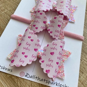 May include: Two pink hair bows with glitter and the words "big sister" and "little sister" printed on them. The bows are on a white background with the text "All that sparkles bows" on the bottom right corner.