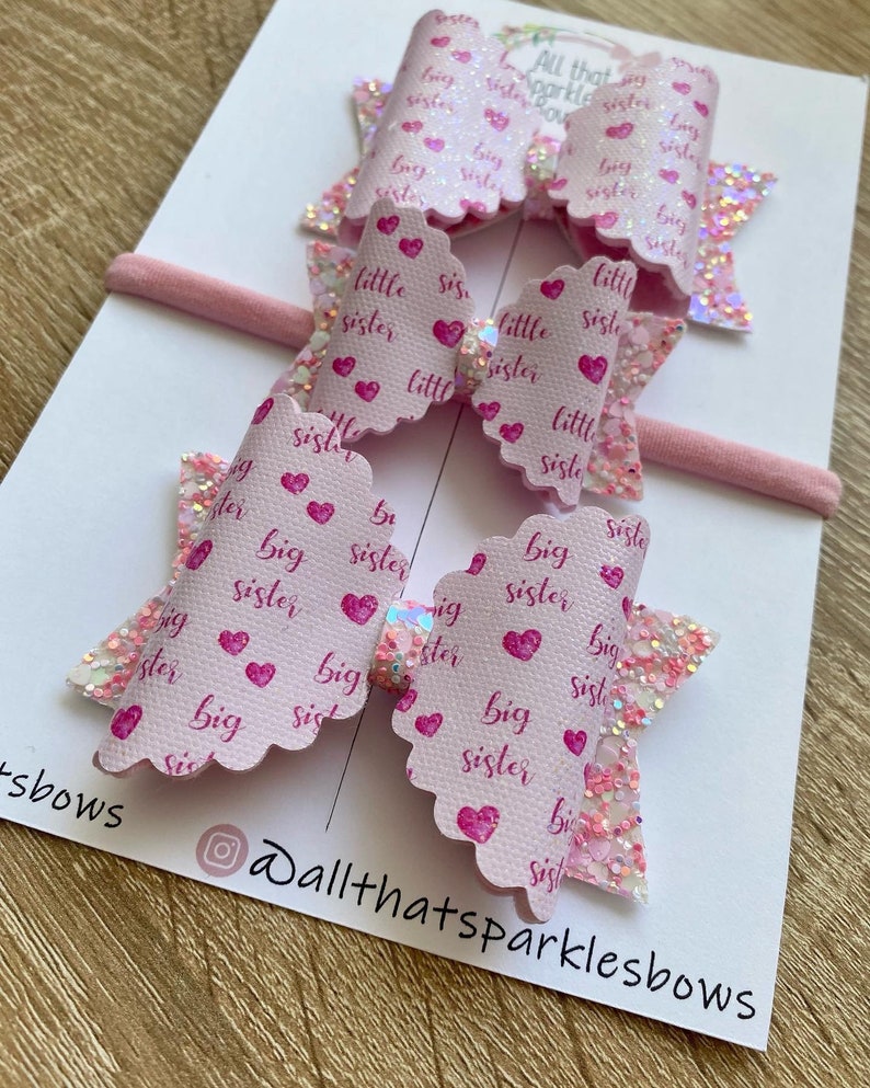 May include: Two pink hair bows with glitter and the words "big sister" and "little sister" printed on them. The bows are on a white background with the text "All that sparkles bows" on the bottom right corner.