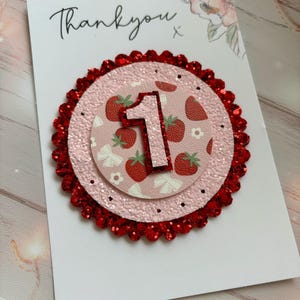 May include: A pink and red glitter card with the number one on it. The number one is surrounded by a strawberry pattern. The card says "Thank you" on the top.