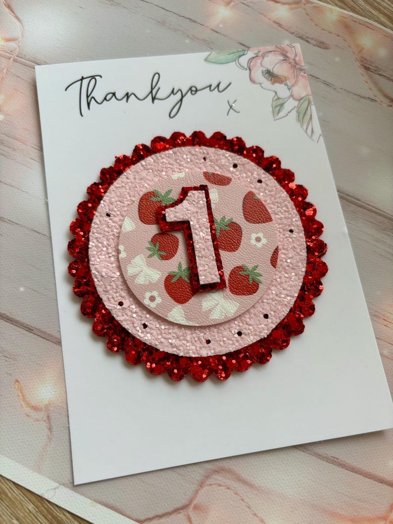 May include: A pink and red glitter card with the number one on it. The number one is surrounded by a strawberry pattern. The card says "Thank you" on the top.
