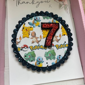 K&ouml;nnte beinhalten: Eine Dankeskarte mit einem runden, mit blauen Glitzer besetzten Rahmen, einer roten Glitzerzahl 7 und einem wei&szlig;en Hintergrund mit einem Pokemon-Muster. Der Text "Danke" steht auf der Karte.