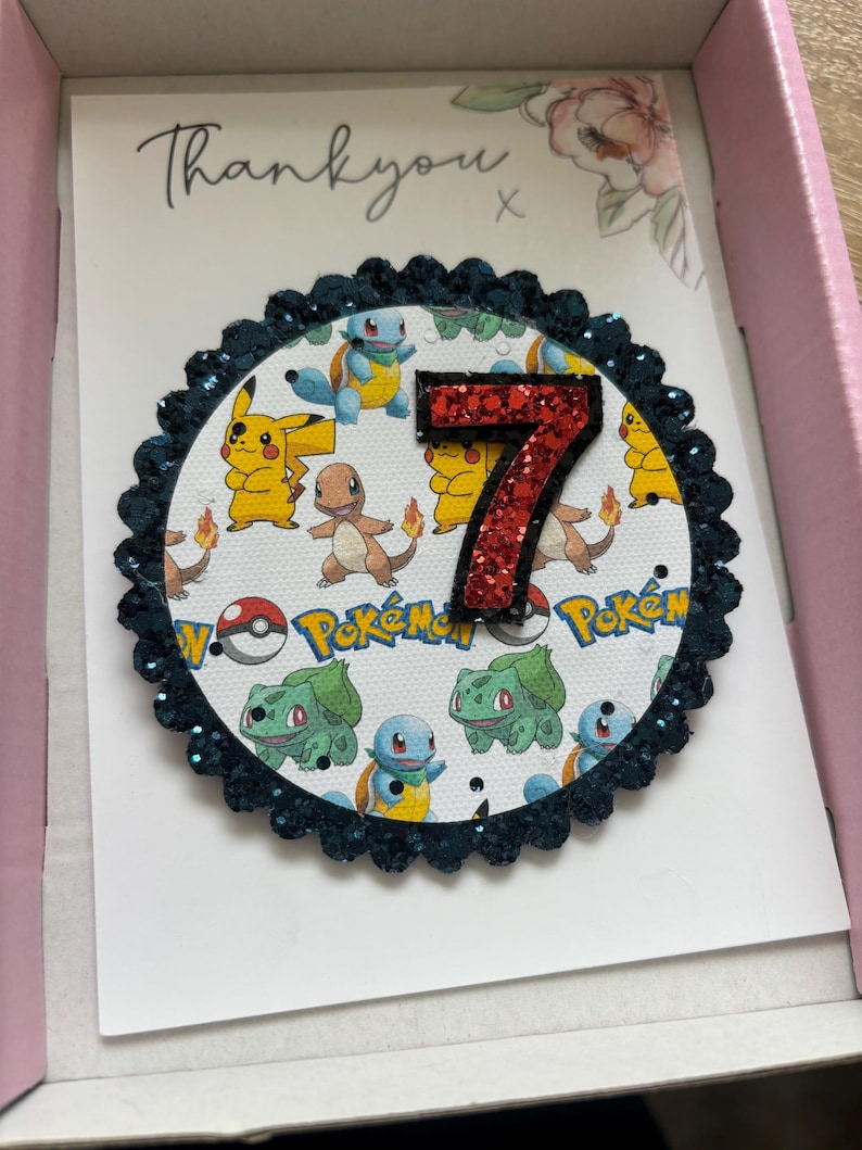 K&ouml;nnte beinhalten: Eine Dankeskarte mit einem runden, mit blauen Glitzer besetzten Rahmen, einer roten Glitzerzahl 7 und einem wei&szlig;en Hintergrund mit einem Pokemon-Muster. Der Text "Danke" steht auf der Karte.