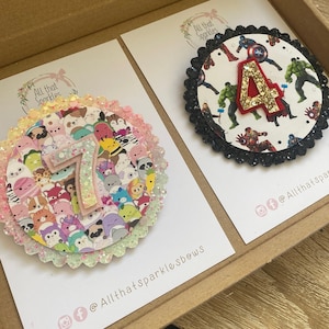 K&ouml;nnte beinhalten: Zwei handgefertigte Filz-Geburtstagsabzeichen mit Glitzerakzenten. Das erste Abzeichen zeigt eine Nummer 7, umgeben von bunten Comic-Tieren. Das zweite Abzeichen zeigt eine Nummer 4, umgeben von Superhelden-Charakteren. Beide Abzeichen haben einen schwarzen gewellten Rand und den Text "All That Sparkles" auf der Karte.