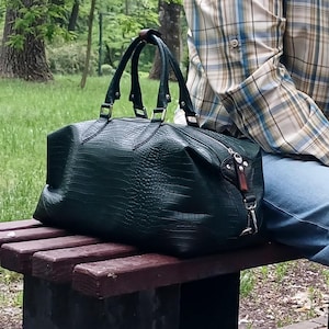 May include: A dark green holdall bag with a textured, reptilian-like embossed pattern. The bag features two top handles, a zippered main compartment, and silver-toned metal hardware, including side clips for an optional shoulder strap attachment.