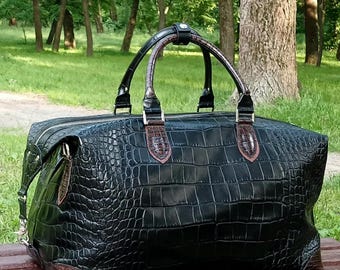 May include: A black duffel bag featuring a textured crocodile skin pattern. The bag includes two dark brown handles with a matching brown leather connector, silver metal hardware, and reinforced brown leather corners at the base.