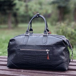 May include: A black duffel bag with a textured crocodile skin pattern. The bag features two top handles, a silver-toned horizontal zipper pocket on the front, and a detachable shoulder strap with silver metal hardware clips on the sides.